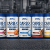 Five containers of Applied Nutrition CARB-X supplement on a dark surface with a blurred gym background.