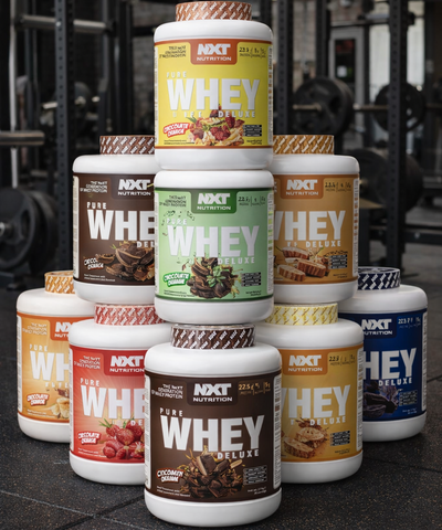 Stack of various flavors of NXT Nutrition Whey protein containers in a gym setting.